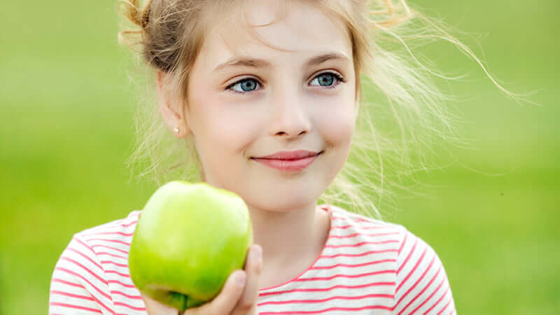 A happy girl holding a green apple, representing the phrase 'Kids They Are What They Eat'.