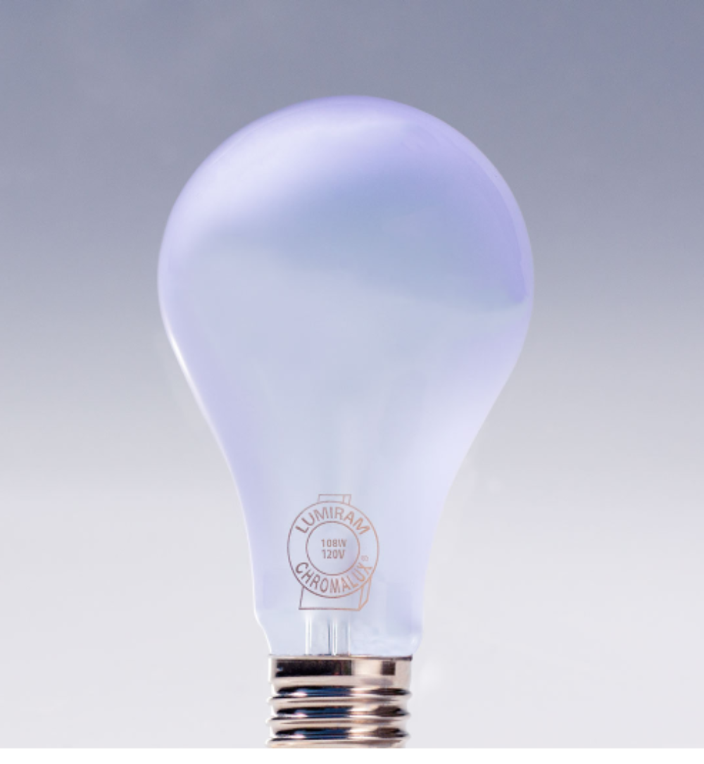A glowing A19-style light bulb with a frosted glass body and a metallic screw base, shown against a soft gray gradient background. The glass has a faint lavender tint from the emitted light, and the base features a circular LUMEN AGE logo.